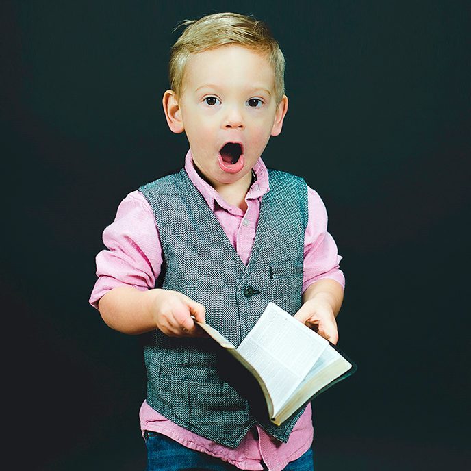 Overrasket barn iklædt lyserød skjorte og grå vest står med en bog i hænderne.
